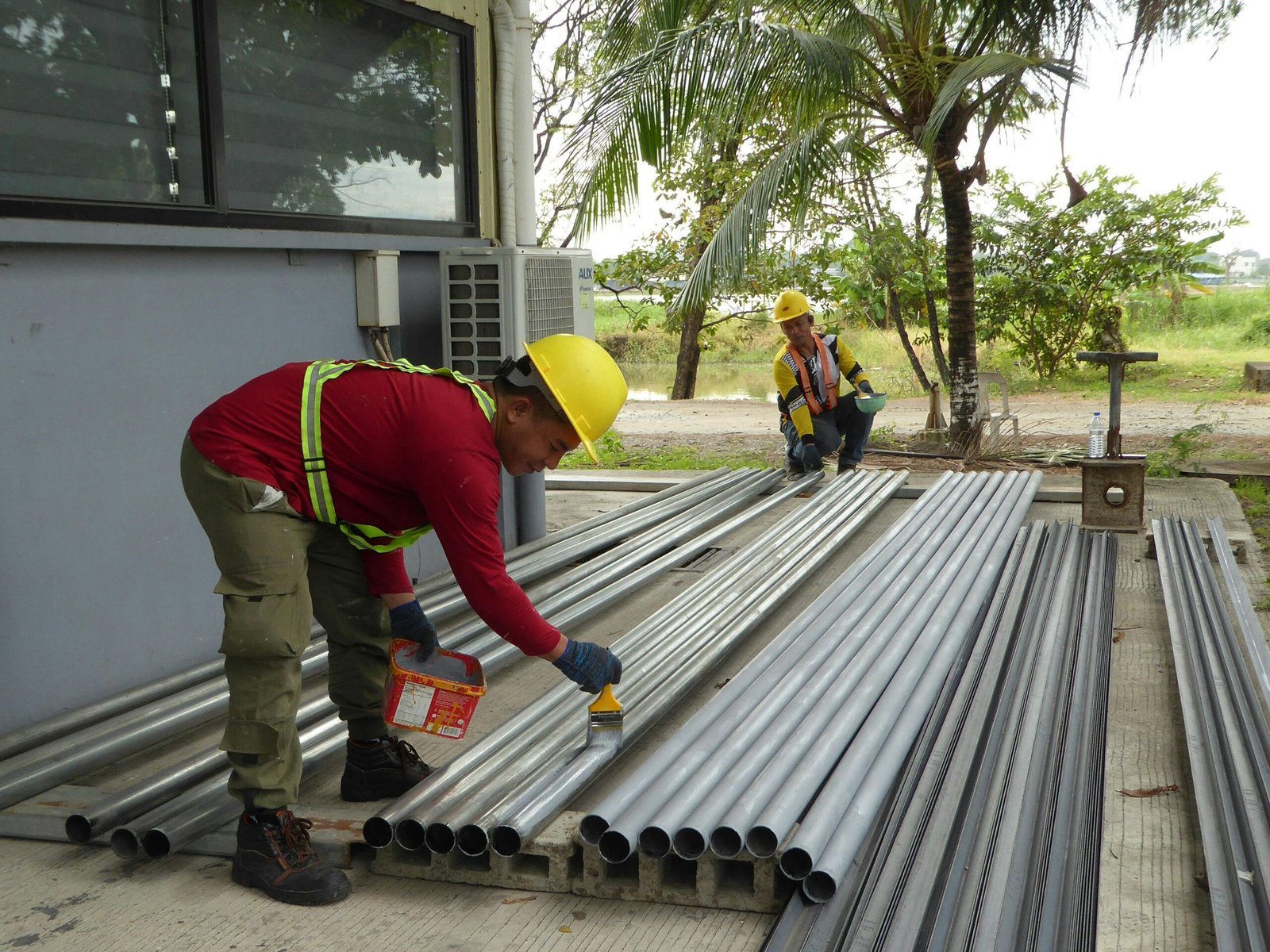 Two construction workers paint metal pipes outdoors, wearing protective gear and helmets.